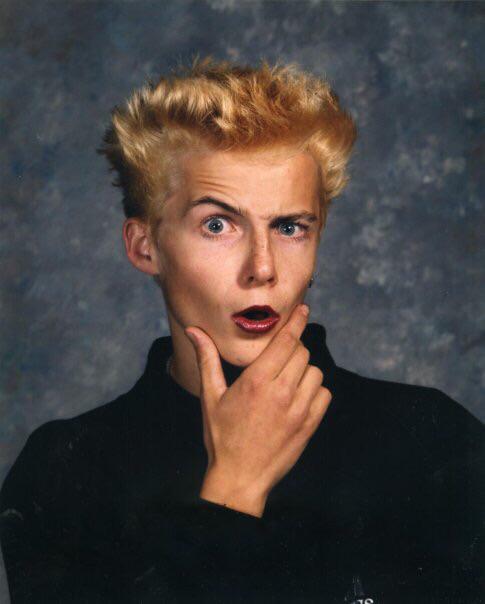 A young person with spiky blond hair and dark lipstick holds their chin and looks surprised, raising one eyebrow. They are wearing a dark shirt and are posed in front of a mottled gray background.