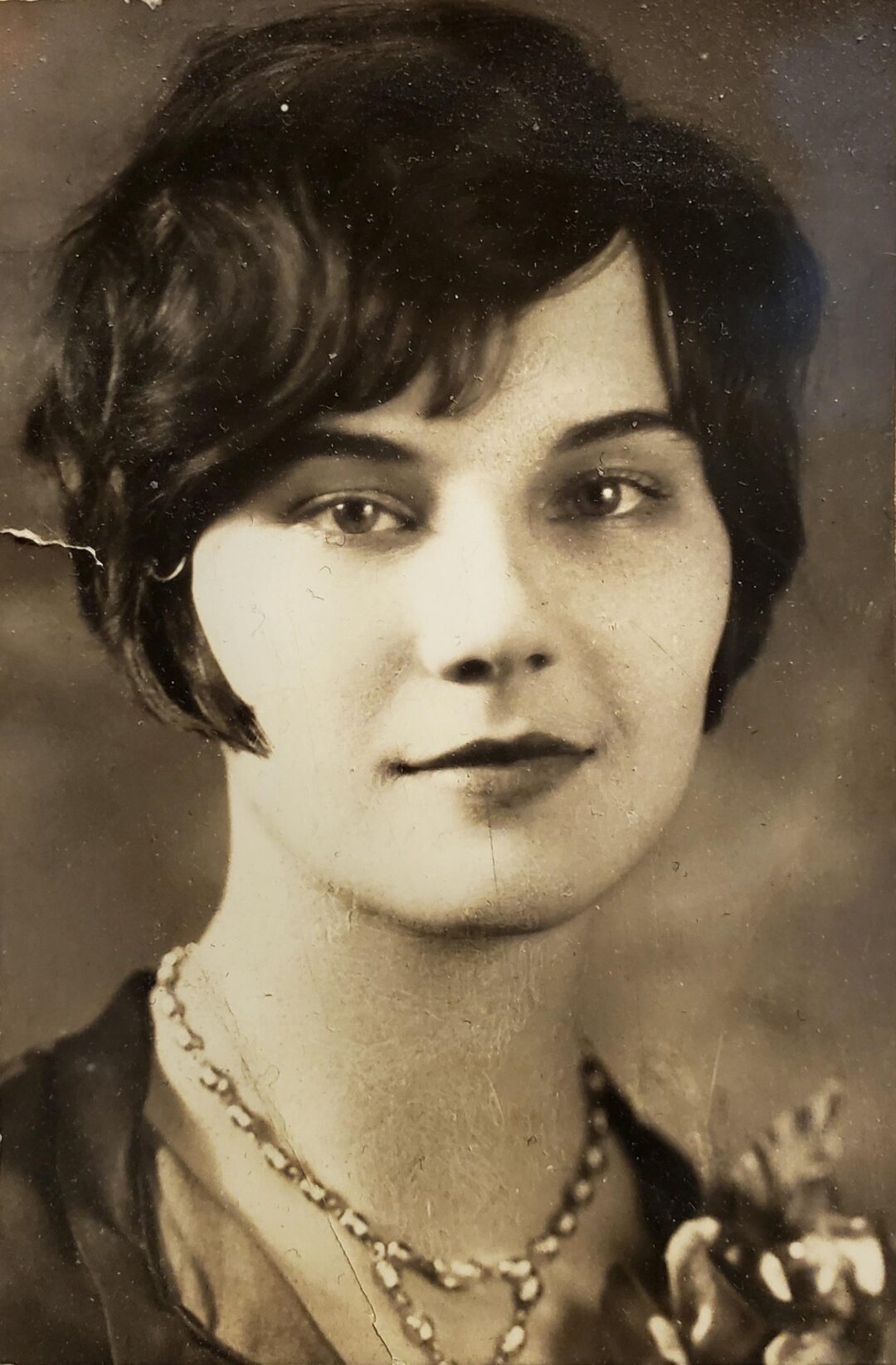 A vintage black-and-white portrait of a young woman with short, wavy dark hair. She is wearing a necklace and a garment adorned with a decorative flower, gazing softly at the camera with a slight smile.