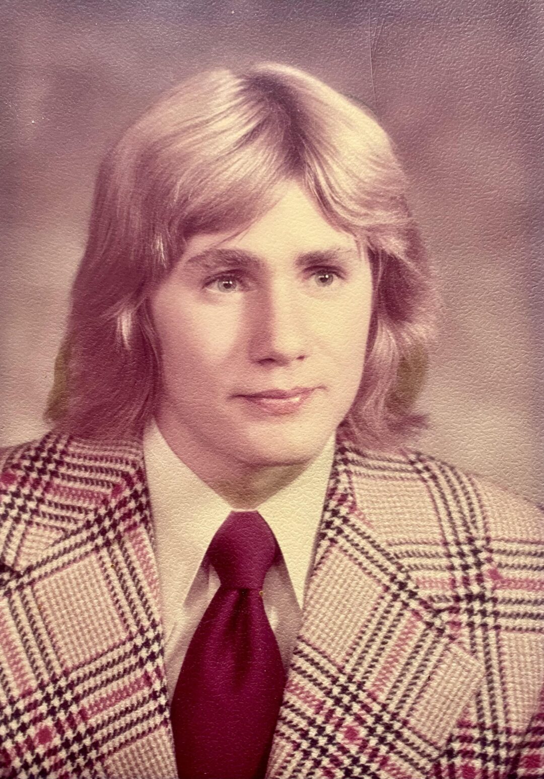 A young person with shoulder-length blond hair wears a patterned plaid jacket, cream shirt, and wide burgundy tie, posing for a formal portrait against a softly blurred studio background.