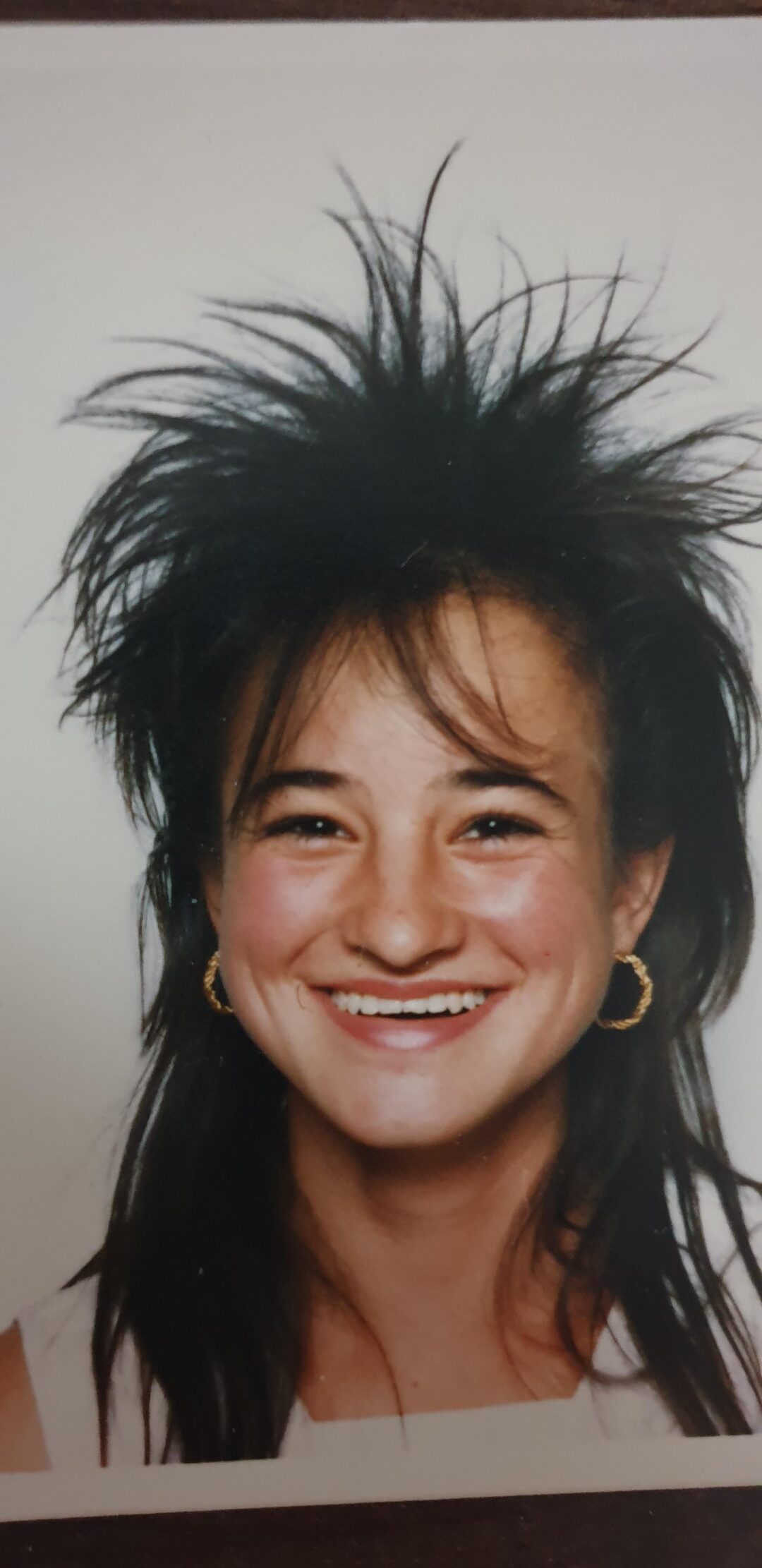 A young woman smiling at the camera, wearing gold hoop earrings and a white top, with voluminous, teased, spiky dark hair styled in an 80s fashion against a plain background.