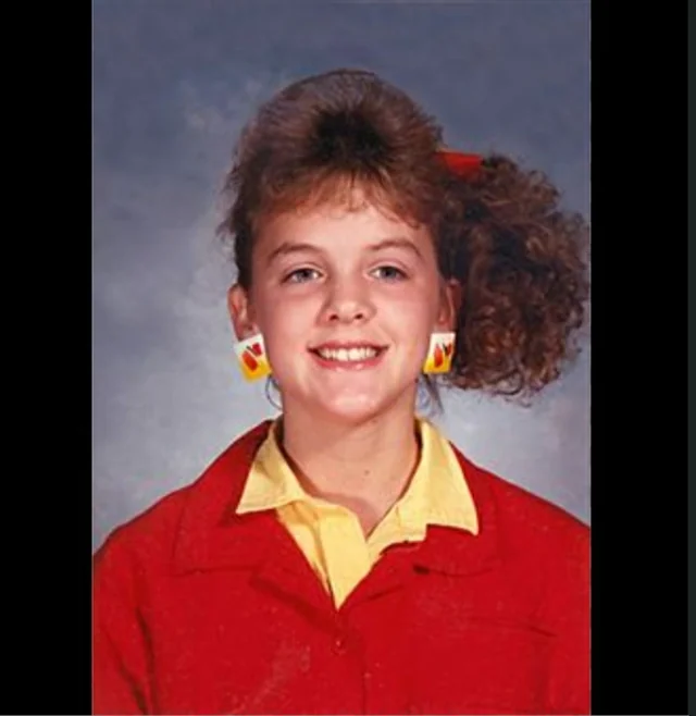 A young girl with curly brown hair tied to the side smiles in a school photo. She wears a red jacket over a yellow shirt and large, colorful geometric earrings. The background is a typical gray school portrait backdrop.