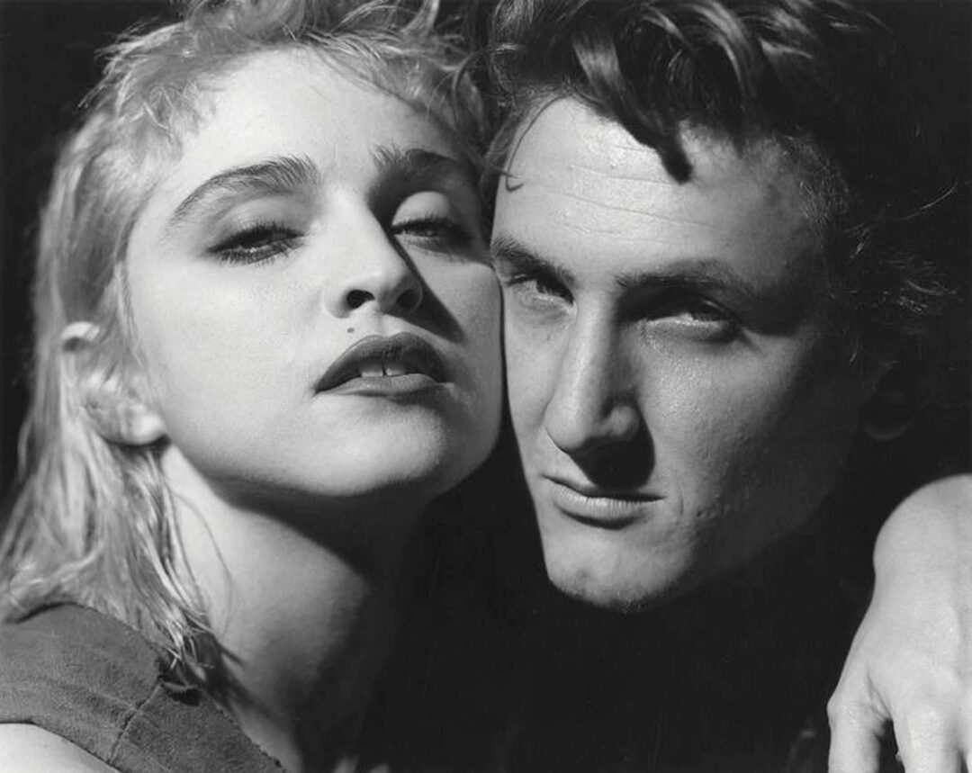 Two people pose closely together in a black and white photo. One has short, light hair and leans toward the camera, while the other, with darker hair, looks intensely forward. Their faces are side by side, showing intimate expressions.