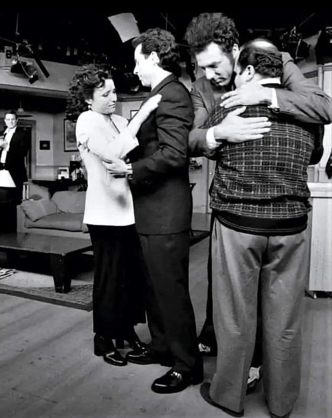 Four people stand in pairs, embracing each other warmly on a TV set. Two women and two men are facing each other. Stage lights and set furniture are visible in the background. The mood appears emotional and nostalgic.