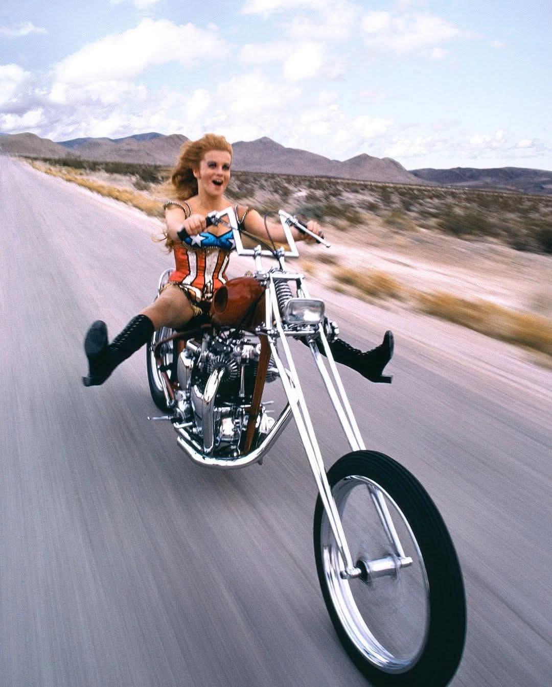 A woman in a corset and boots rides a custom chopper motorcycle fast on a desert road, smiling with her legs extended forward and her hair blowing in the wind. Mountains and blue sky are in the background.