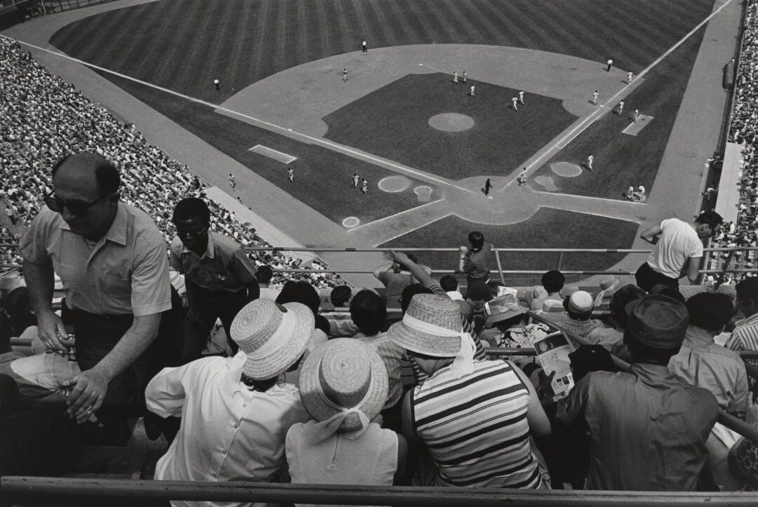 A crowded baseball stadium seen from the stands behind home plate. Fans in hats sit close together, watching players on the field below during a daytime game.