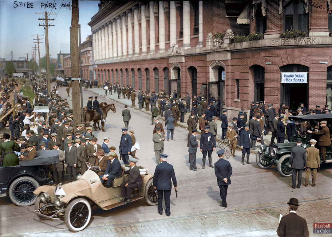 Crowd of people in early 20th-century attire gather outside Shibe Park baseball stadium. Vintage cars, a horse and carriage, and streetcars are visible along the busy street, with a brick building in the background.