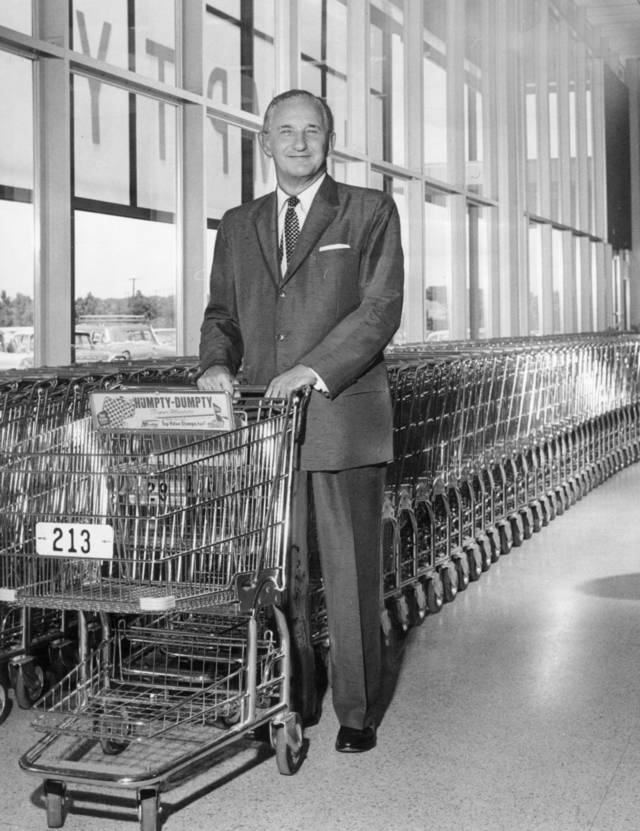 A man in a suit stands inside a supermarket, smiling and holding the handle of a shopping cart labeled "213" in front of a long row of nested shopping carts by large windows.