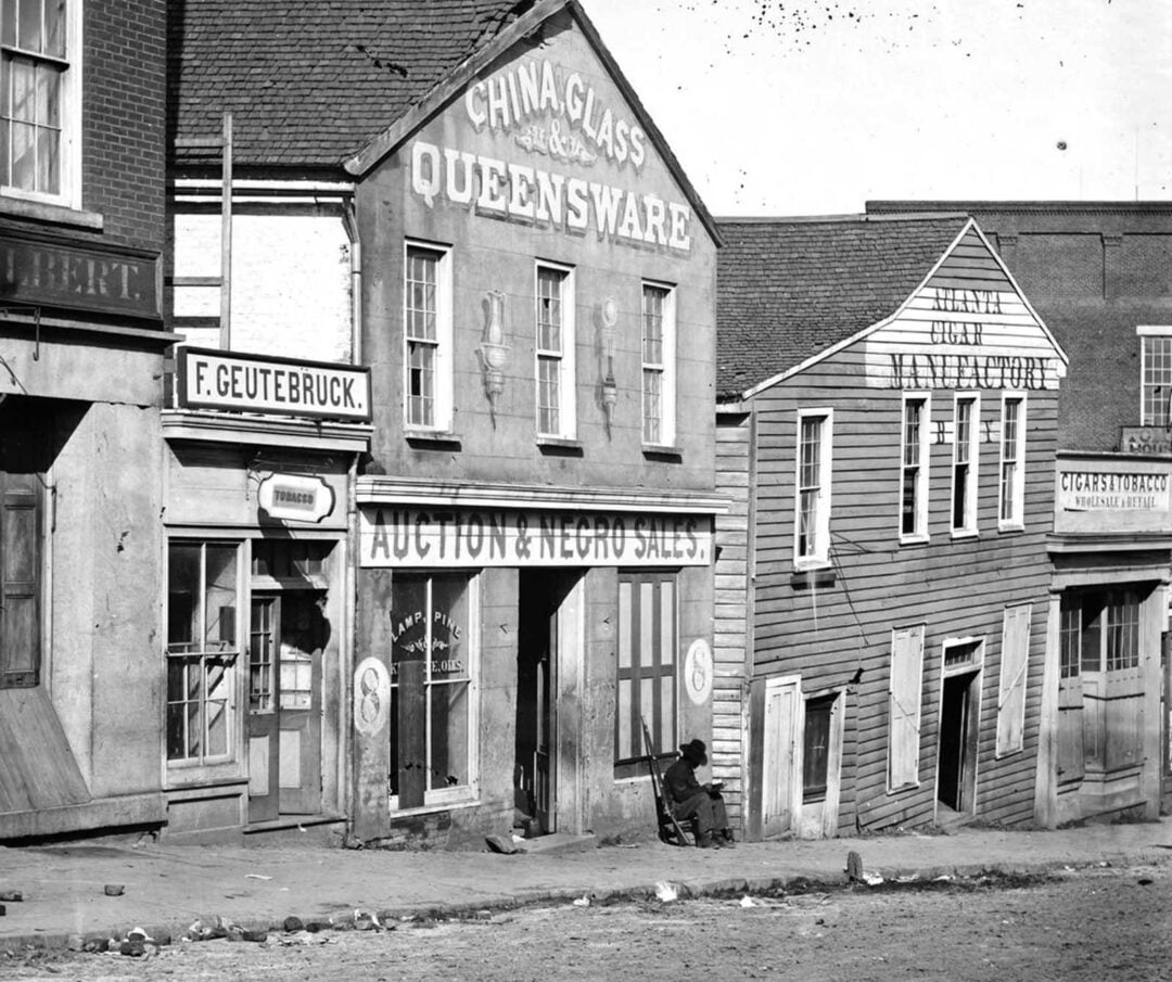 Black-and-white photo of old storefronts, including signs for "China, Glass, Queensware," "Auction & Negro Sales," and others. A person sits on the sidewalk outside the buildings on a sloped dirt street.