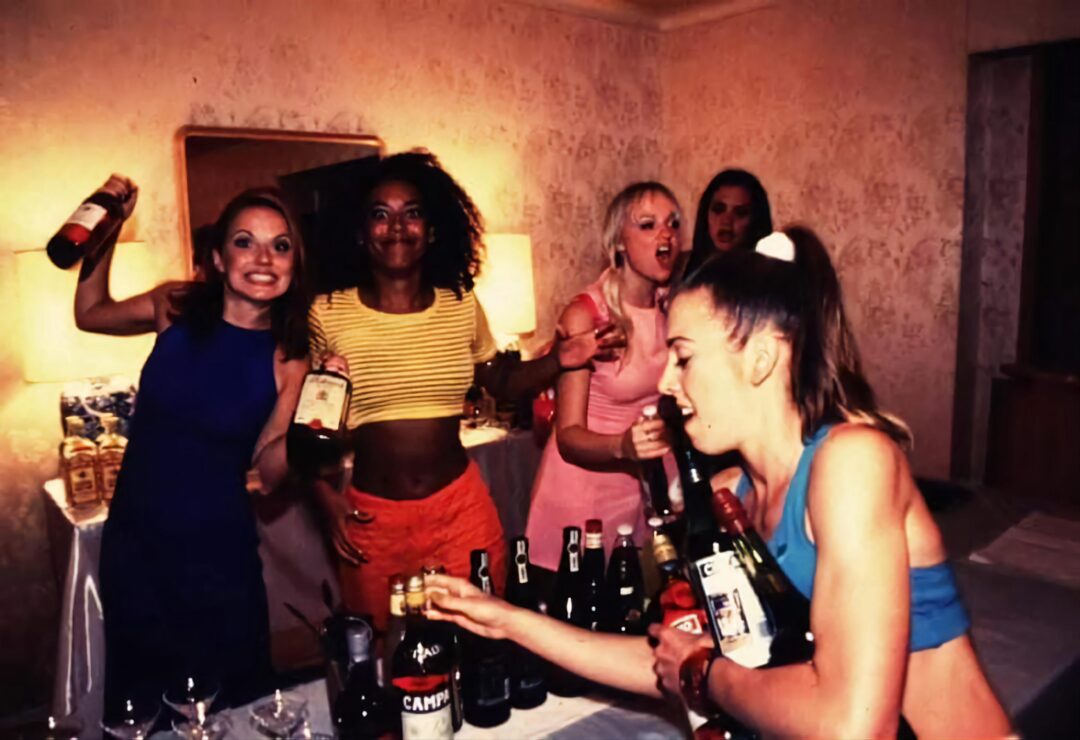 Five women in colorful outfits stand in a room, laughing and holding bottles of alcohol, with more bottles on the table in front of them. The background shows a light wallpaper and a mirror on the wall.