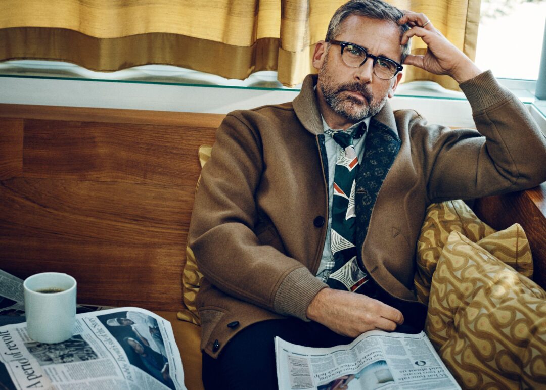 A man with glasses and a beard sits on a couch, resting his head on one hand. He wears a brown jacket and patterned tie, with newspapers and a cup of coffee in front of him. Yellow curtains hang behind him.