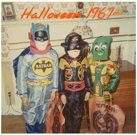 Three children in Halloween costumes—Batman, a cowboy, and Gumby—stand indoors holding orange trick-or-treat bags. "Halloween 1967" is written in orange at the top of the photo.