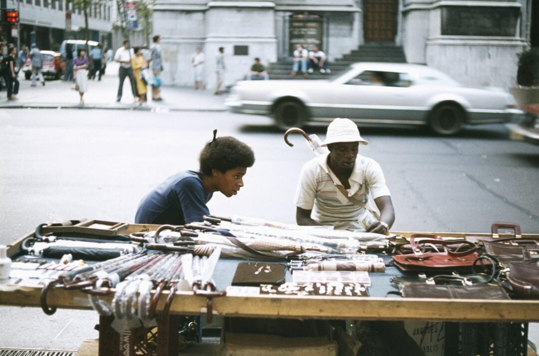 Two street vendors sit at a table displaying umbrellas, belts, and small items on a city sidewalk. A car drives by in the background, and people sit on steps across the street.