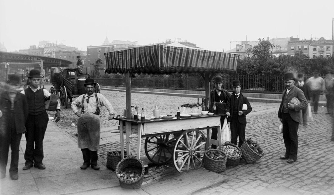 Black and white photo of men standing around a street vendor cart filled with baskets of produce on a cobblestone road, with buildings and trees in the background. One man wears an apron; others are dressed in suits and hats.