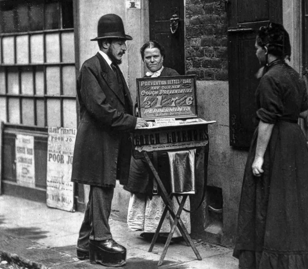 A man in a suit and bowler hat stands at a street stall labeled “Cough Prevention,” speaking to two women on a narrow, cobbled street beside brick buildings in an old, urban setting.