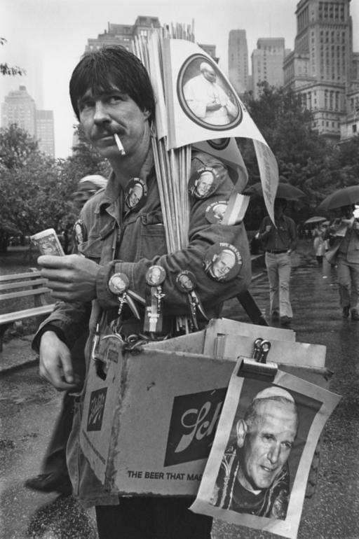 A man stands outdoors holding religious pins and posters, with buttons and a Pope photo attached to his jacket and a cardboard box. City buildings and people with umbrellas are in the background.