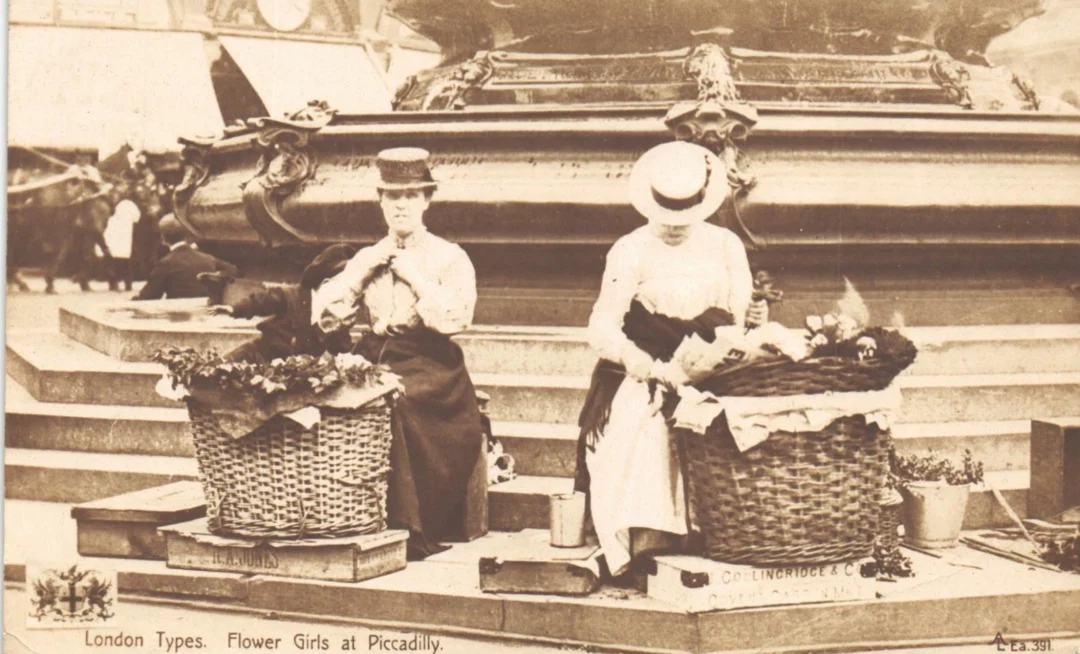 Two women in Edwardian-era clothing sit beside large baskets of flowers near a monument in Piccadilly, London. Both wear hats and are arranging flowers, with city life blurred in the background.