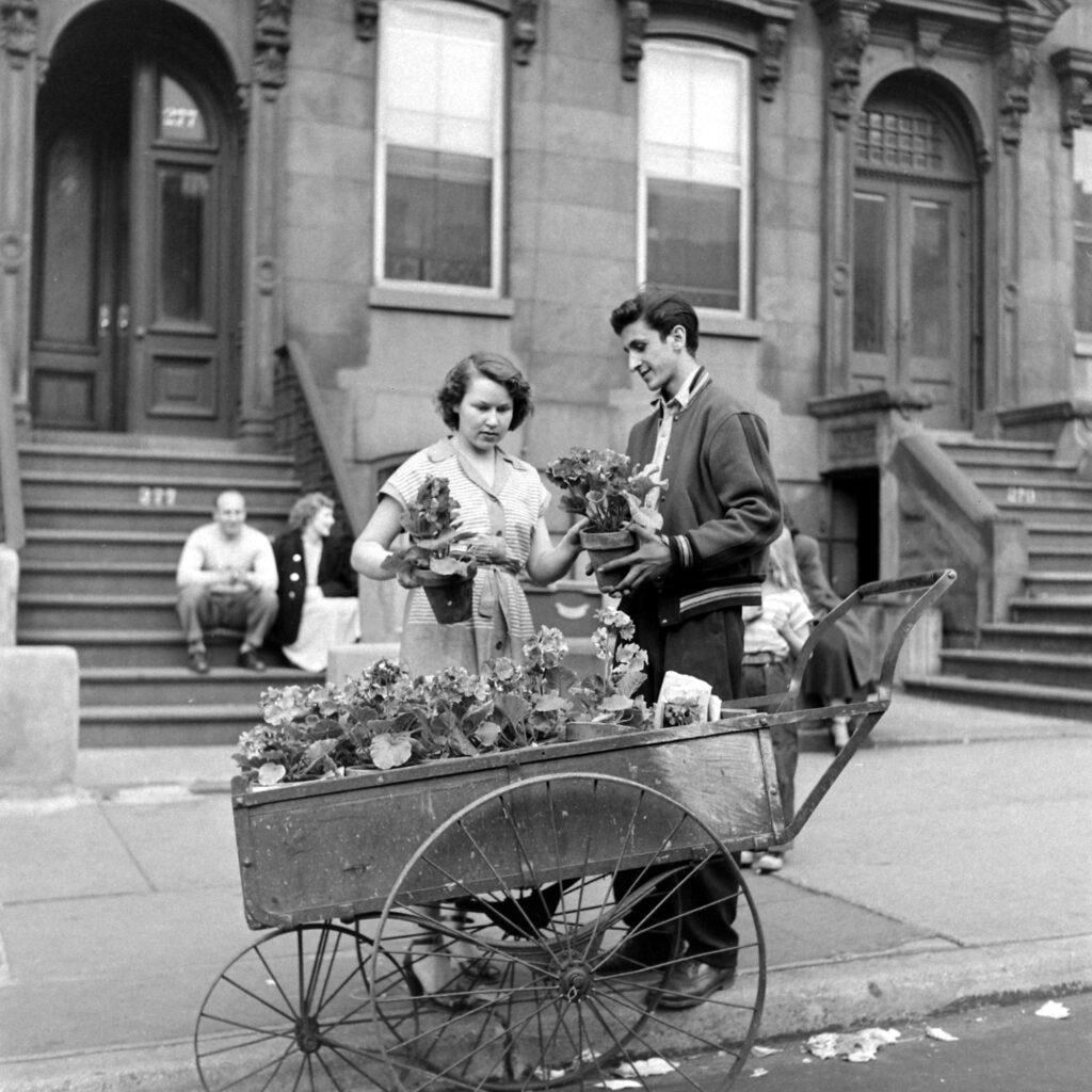 A young woman and man stand beside a wooden cart filled with potted flowers on a city sidewalk. Stone building stoops and seated people appear in the background. The scene looks vintage, likely from the mid-20th century.