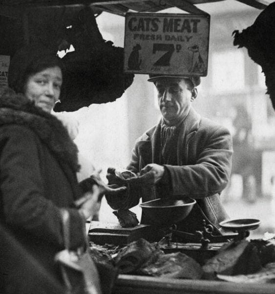 A man behind a market stall weighs meat while a woman stands in front. A sign above reads “Cats Meat Fresh Daily.” The scene is in black and white, suggesting an old historical marketplace.