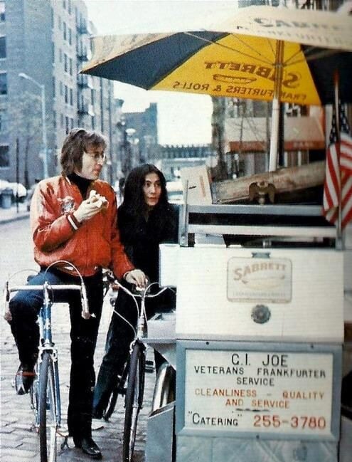 A man in a red jacket and a woman stand by a street hot dog cart with "C.I. JOE Veterans Frankfurter Service" signage. The man holds a sandwich and sits on a bicycle; city buildings are visible in the background.