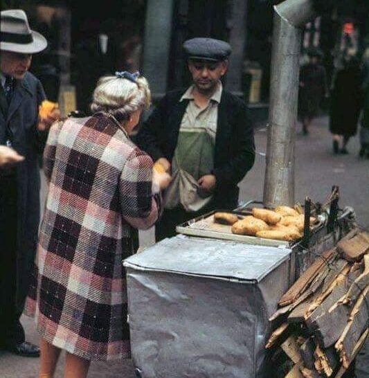 A street vendor wearing a cap sells baked goods from a cart with a metal oven and firewood on a busy city sidewalk, as two customers, one in a plaid coat, make a purchase.