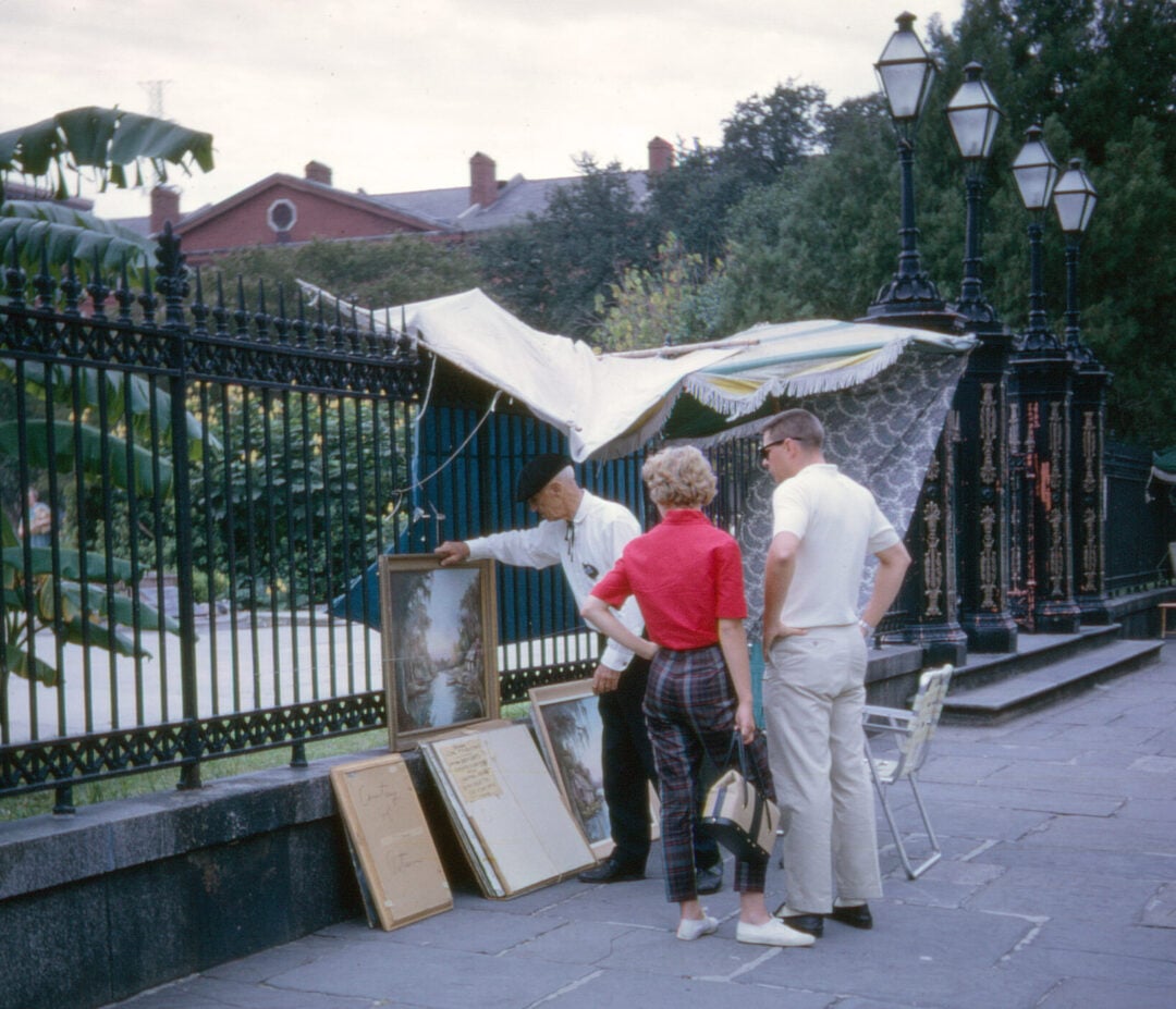 A street artist shows framed paintings to a woman and a man on a sidewalk by an iron fence, with art displayed on the ground and lamps and trees in the background.
