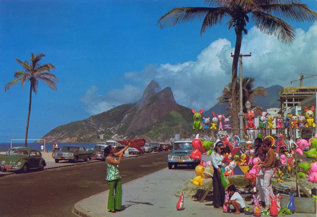 A vibrant street scene by the beach with palm trees, colorful inflatable toys, vintage cars, and people shopping. Mountains and blue sky are visible in the background.