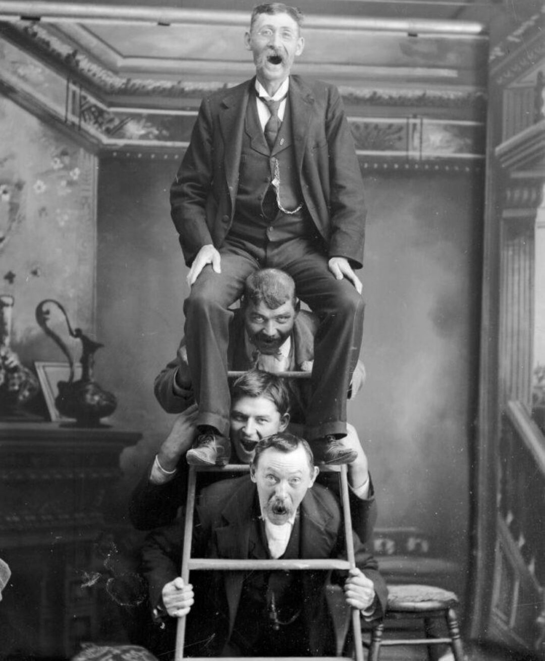 Four men in suits pose in a humorous human pyramid, each sitting on the shoulders of the one below, with surprised facial expressions in a vintage, indoor setting with elaborate decor in the background.