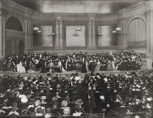 A large crowd in formal attire gathers in a grand concert hall with ornate walls and an organ. Many people are seated onstage and in the audience, suggesting a formal event or ceremony in the early 20th century.
