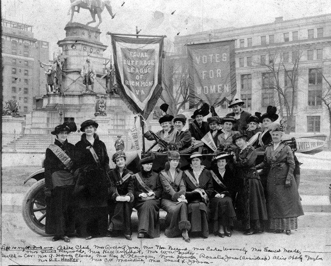 A group of women suffragists in early 1900s attire pose in front of a monument, holding banners that read “Votes for Women” and “Equal Suffrage League of Richmond.” Some wear sashes and hats; buildings are visible in the background.