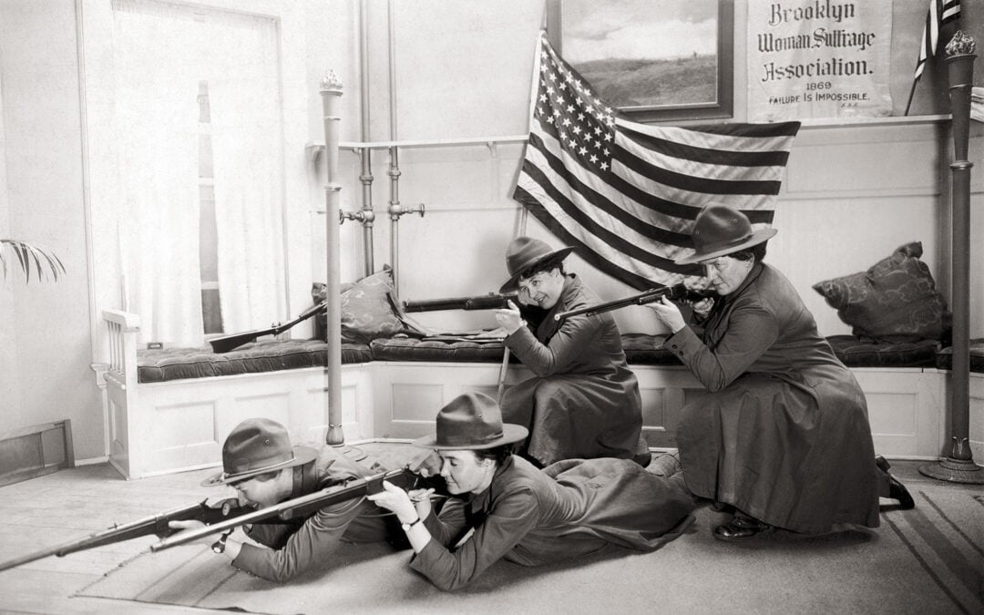 Four women in long dresses and hats pose with rifles in a room, an American flag behind them. A sign reads "Brooklyn Woman Suffrage Association." The women appear determined, emphasizing their activism.