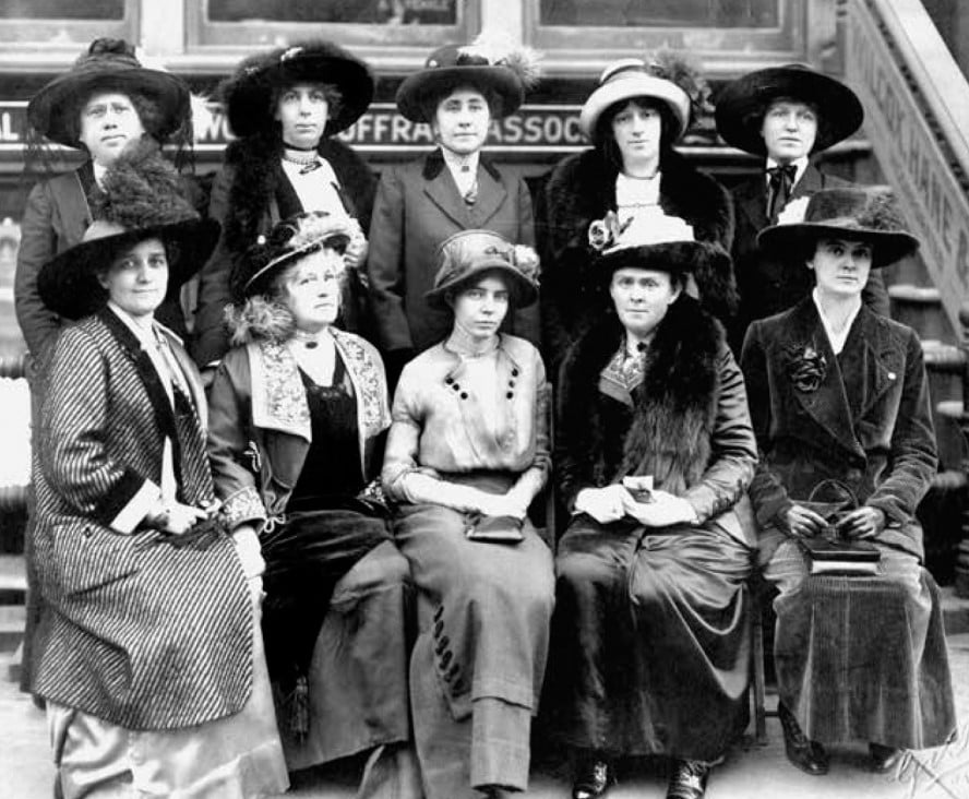 A black-and-white photo of eleven women in early 1900s attire and hats, posed in front of a building with a sign that reads “WOMAN SUFFRAGE ASSOC.” The women are seated and standing in two rows.