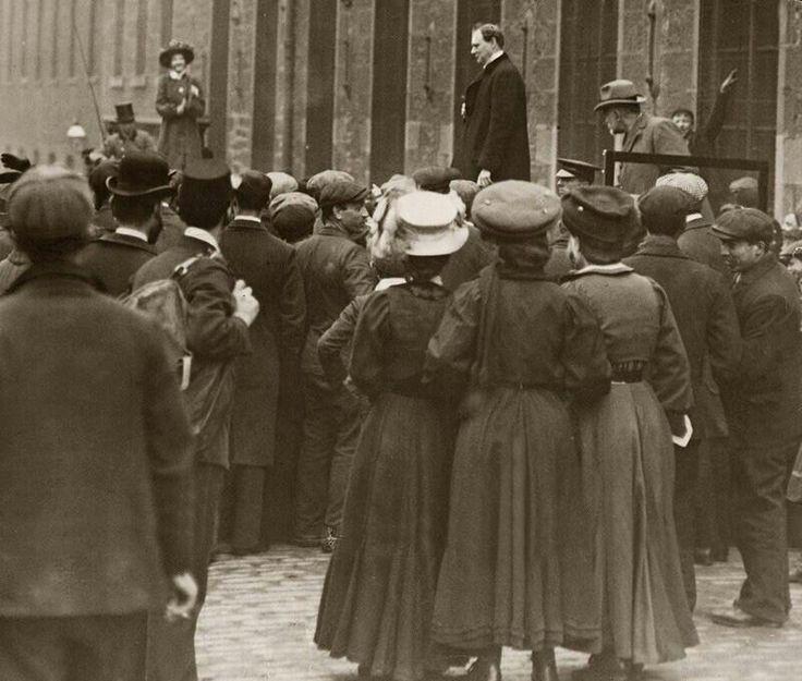 A black-and-white photo shows a crowd gathered around a man standing on a platform, speaking outdoors. Most people wear early 20th-century clothing, including hats and long coats. Two women stand apart, watching from a distance.