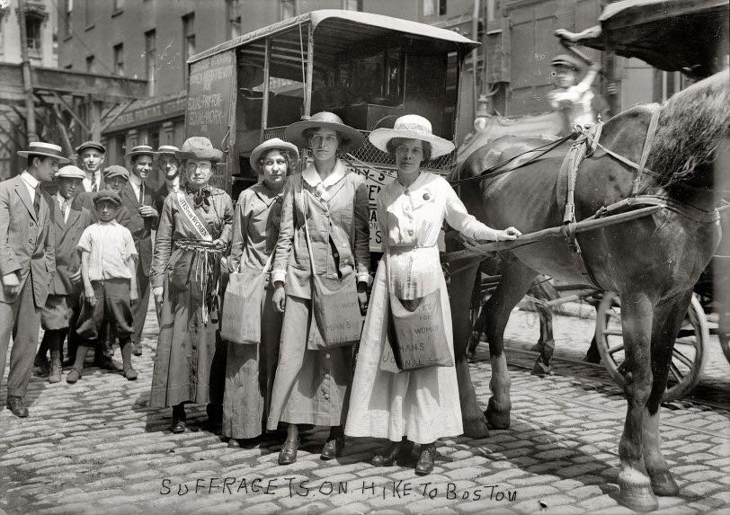 A group of women suffragists wearing hats and sashes stand in front of a horse-drawn wagon on a cobblestone street, surrounded by onlookers. The image is labeled "Suffragists on Hike to Boston.