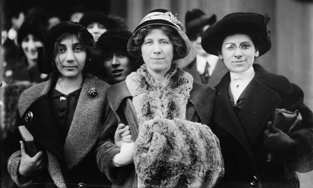 A black-and-white photo of three women in early 20th-century clothing and hats, standing close together and facing the camera, with more people visible in the background. The woman in the center wears a fur stole.