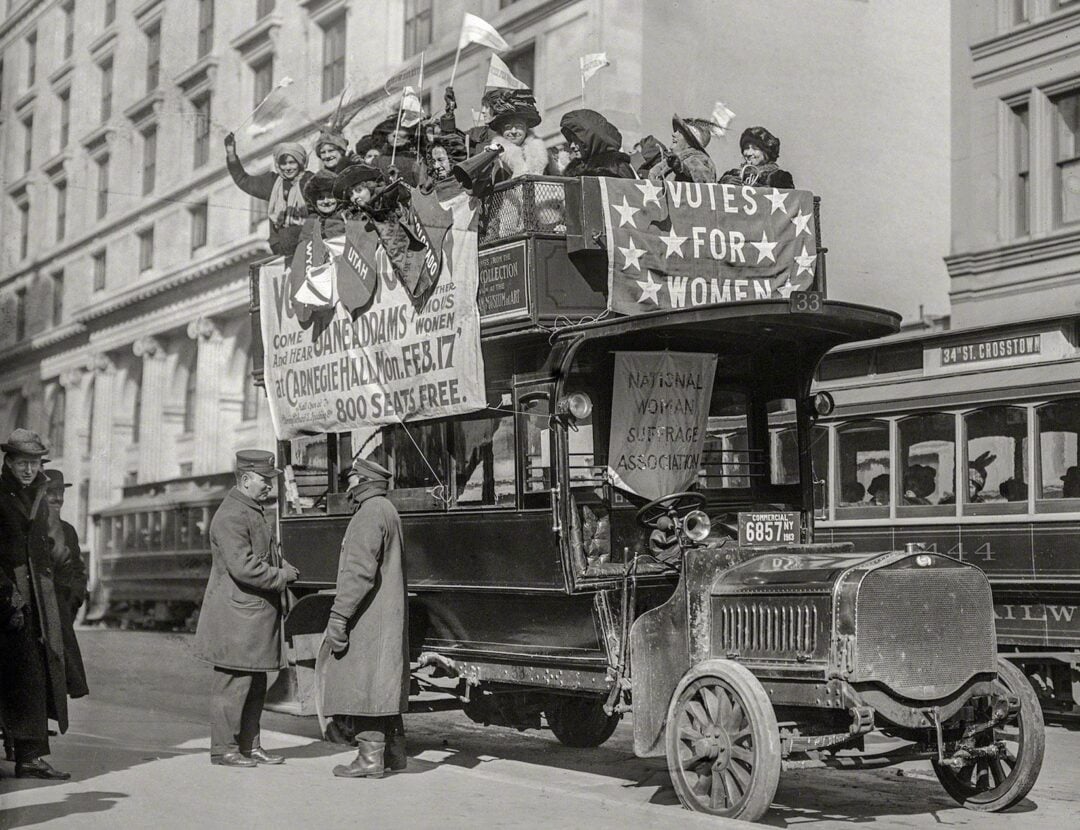 A black-and-white photo of women on a double-decker bus holding flags and banners that read "Votes for Women" during a suffrage parade. Several women wave from the top, and people stand around the bus on a city street.