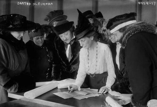 A group of women, including Bertha M. Furman, gather around a table looking at documents. Furman points to the papers, teaching the group. The women are dressed in early 20th-century clothing and hats.