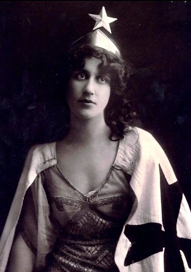 A woman with wavy dark hair wears a star-topped headpiece and a draped garment decorated with a large star. She gazes softly at the camera against a dark background. The image is in black and white.