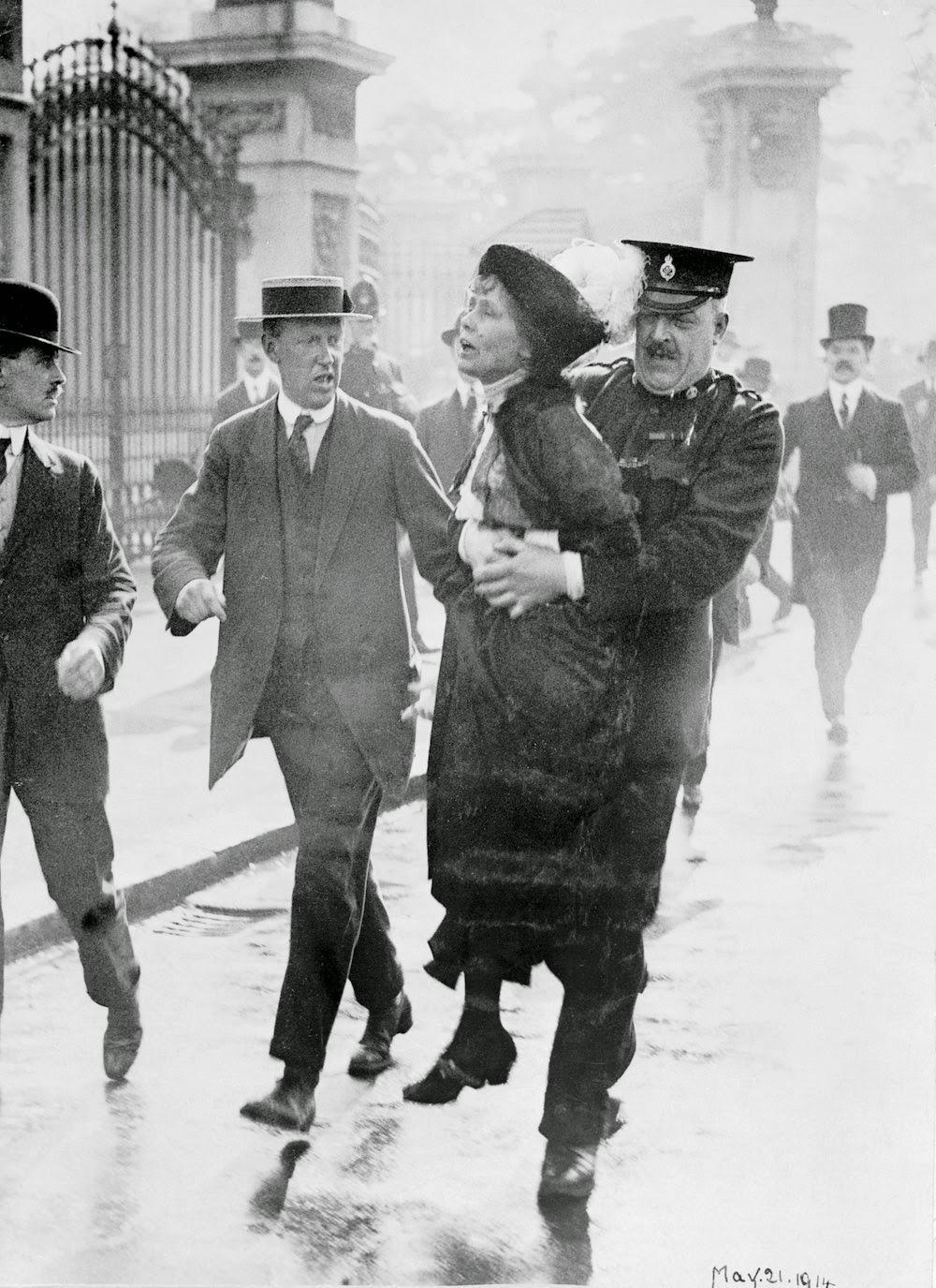 A black-and-white photo shows a police officer arresting a woman while two men in suits walk beside them. The woman appears defiant as she is carried away, with onlookers and tall gates in the background.