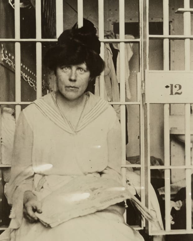 A woman in early 20th-century clothing sits in front of jail cell bars, holding papers in her hands and looking toward the camera. The cell door is marked with the number 12.