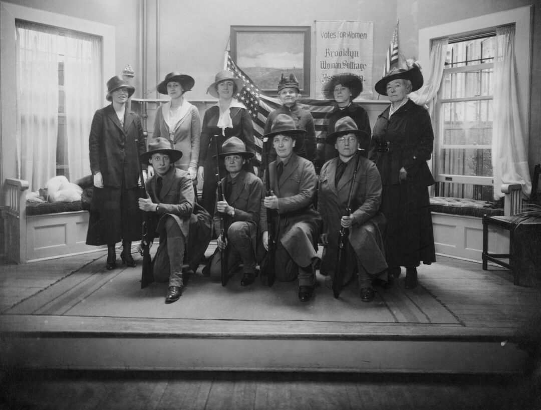 A group of women, some standing and some kneeling with rifles, pose indoors near American flags and a “Votes for Women” sign, wearing early 20th-century clothing and hats, in a black-and-white photo.