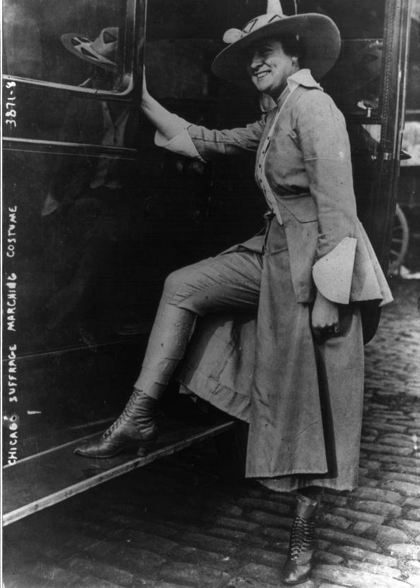 A smiling woman wearing a large hat, long coat, and matching trousers poses with one foot on a running board beside a vehicle on a cobblestone street. Handwritten text reads “Chicago Suffrage Marching Costume.”