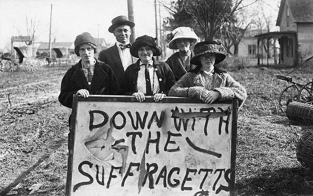 Five people, including young girls and a man, stand outdoors behind a large hand-painted sign reading "DOWN WITH THE SUFFRAGETTS." The group appears to be in a rural area with houses and trees in the background.