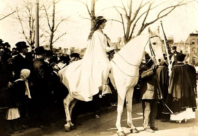 A woman in a flowing white garment sits side-saddle on a white horse during a parade, surrounded by a crowd of people wearing early 20th-century clothing. Leafless trees and buildings are visible in the background.