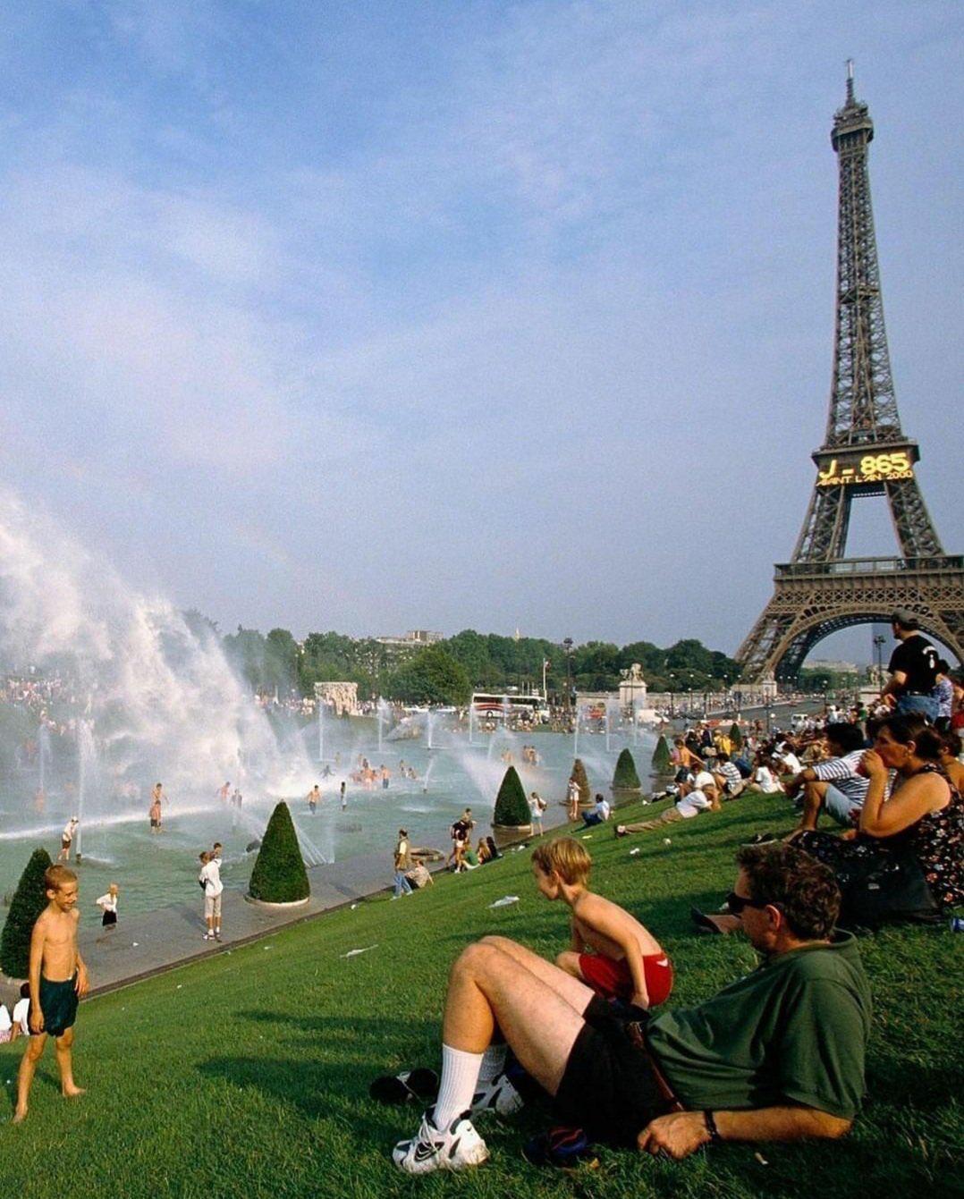 People relax on a grassy slope near the Eiffel Tower, with children and adults playing in water fountains on a sunny day in Paris. The scene is lively and the sky is clear.