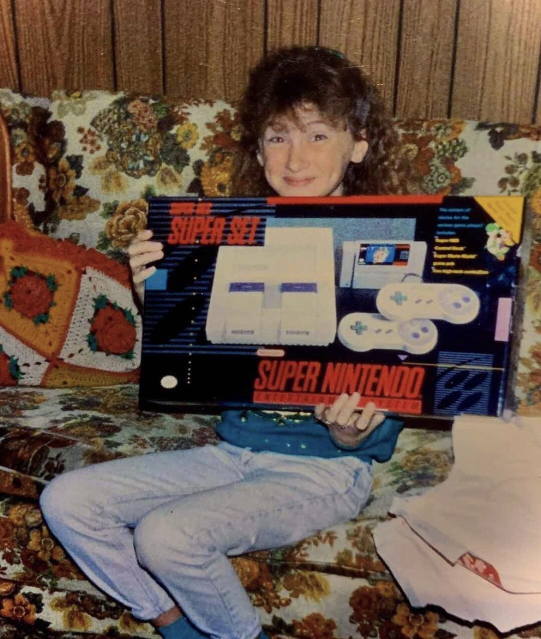 A smiling person sits on a patterned couch holding a boxed Super Nintendo Entertainment System. A crocheted cushion and wood-paneled wall are in the background.