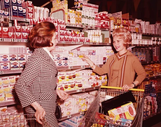Two women in vintage clothing talk and smile in a grocery store aisle, one pushing a shopping cart filled with groceries while the other points at boxed products on the shelves. The scene appears to be from the 1960s.