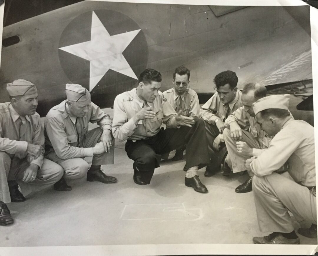 Seven men in military uniforms crouch and sit in a semicircle on the ground, discussing plans sketched in chalk. They are in front of a large aircraft displaying a star insignia.