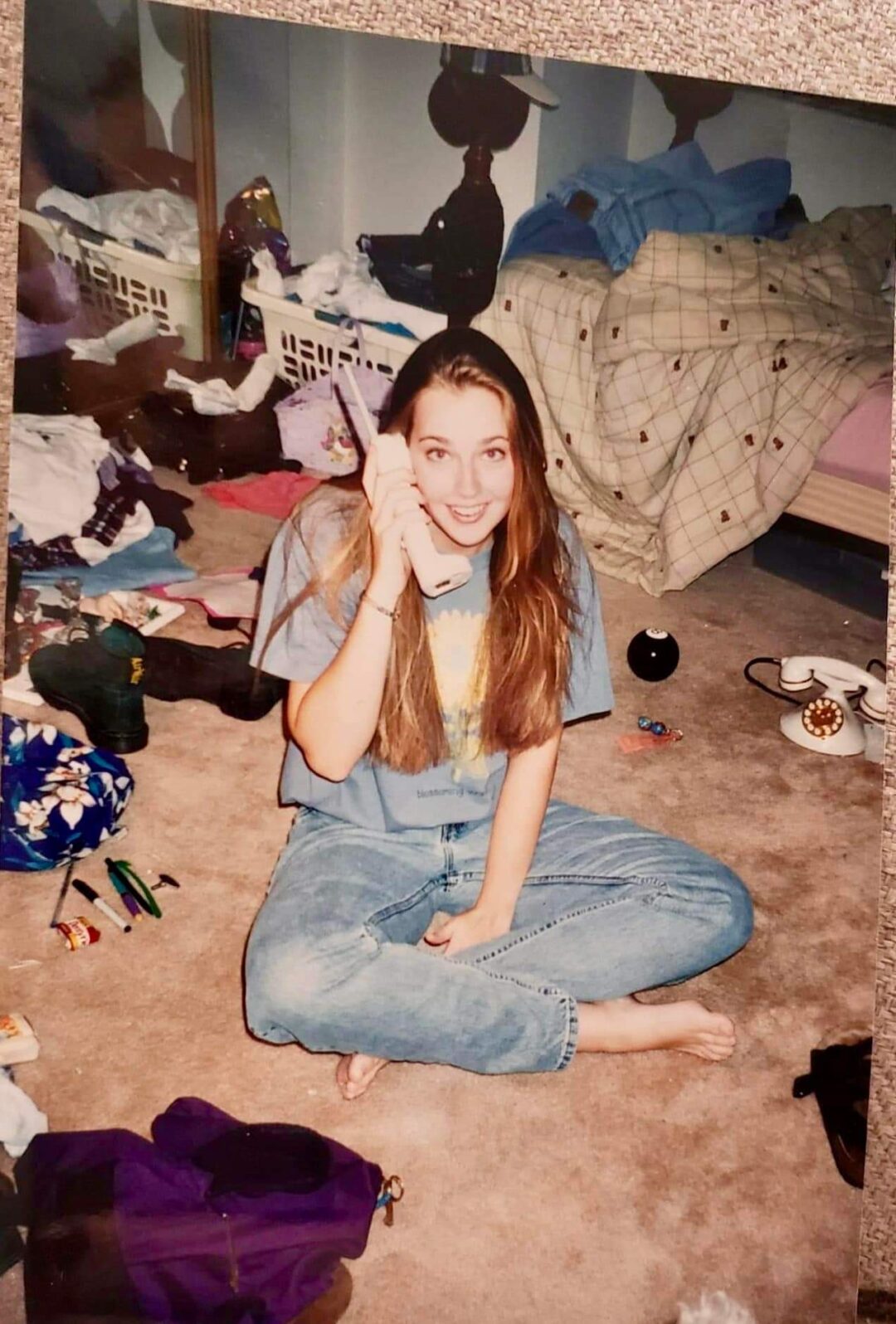 A young woman with long hair sits cross-legged on a messy bedroom floor, smiling while holding a corded phone to her ear. The room has scattered clothes, a cluttered dresser, and an unmade bed in the background.