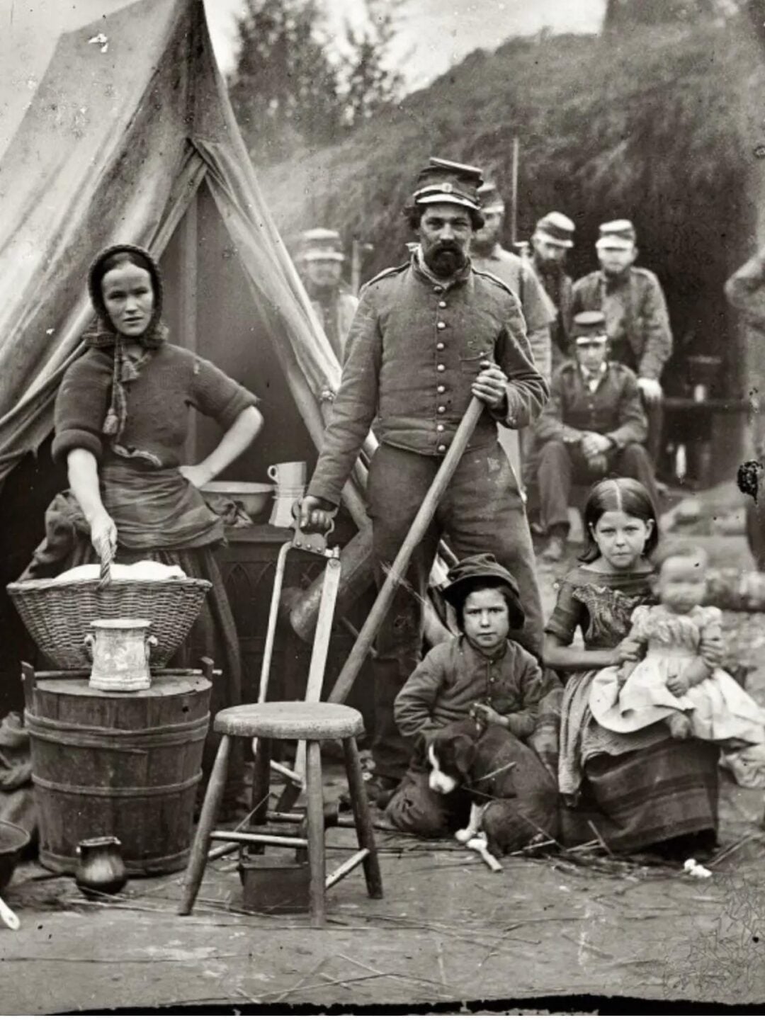 A black-and-white photo of a 19th-century military family outside a canvas tent: a soldier stands with an axe, a woman sits by a basket, and three children sit on the ground, with uniformed men in the background.