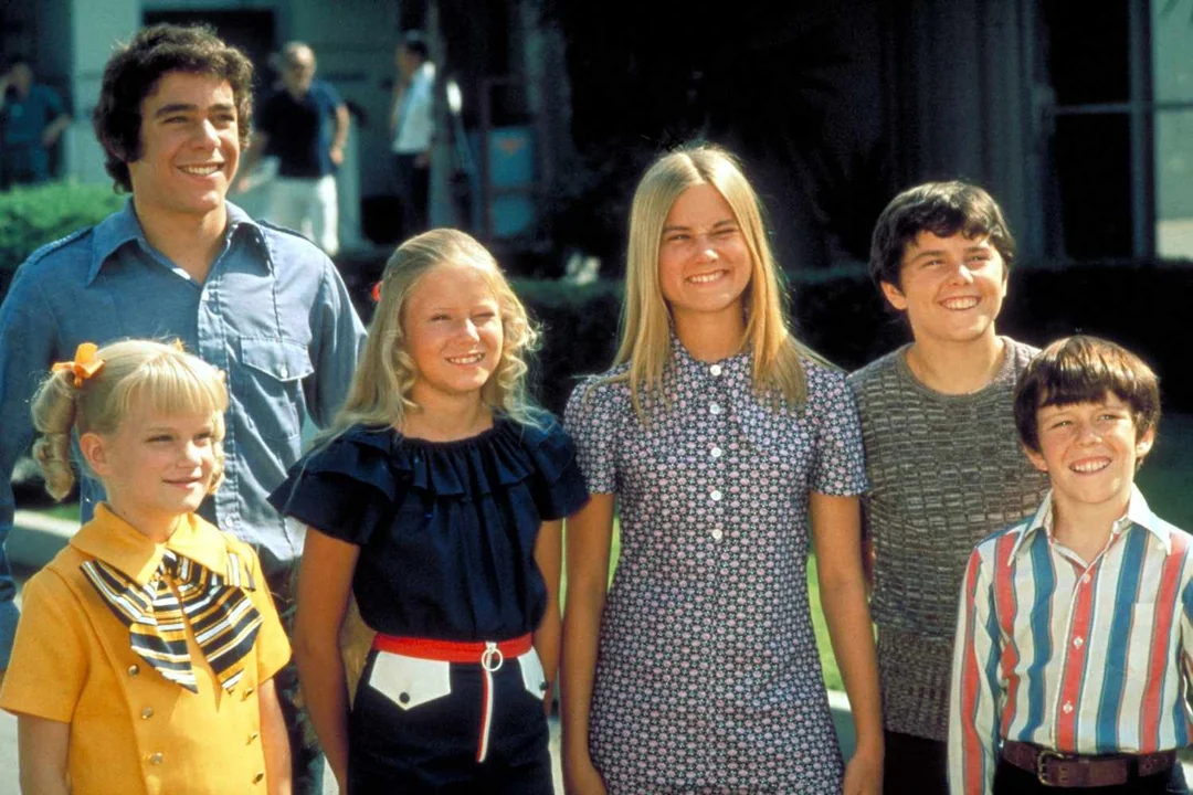 Six smiling children and teenagers stand together outdoors in colorful 1970s clothing, posing for a photo with greenery and a building in the background.